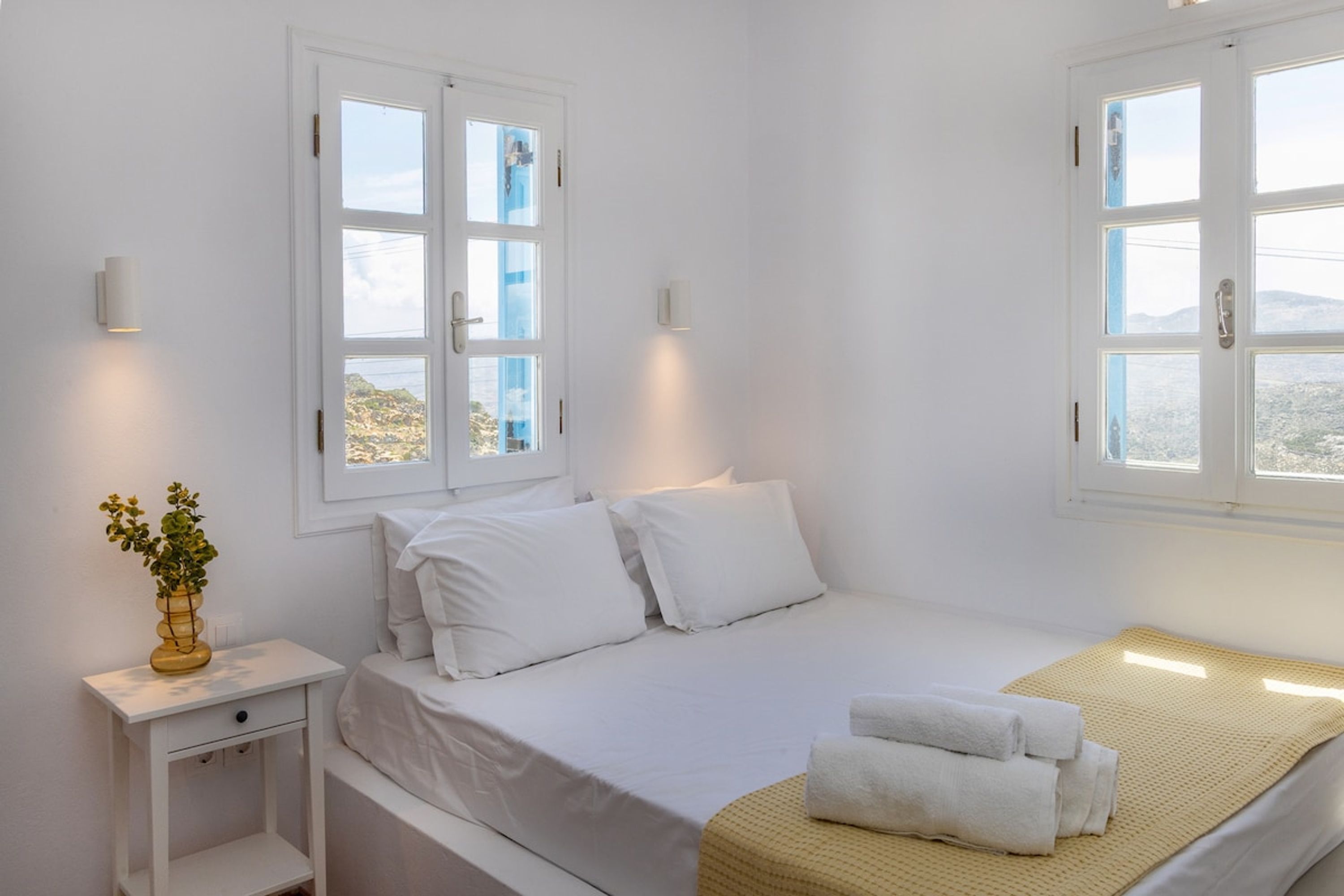 A white room with a bed, two windows, and a bedside table with a vase.