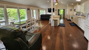 A spacious living area with a leather sofa, a dining table with chairs, a kitchen with white cabinets, and a large window offering a view of greenery. - Zenith at Copalis Rocks - Unsurpassed Ocean View with new Hot Tub (Copalis Beach)