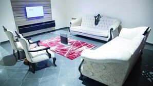 A modern living room with a white sofa, two armchairs, a glass coffee table, and a red patterned rug. A flat-screen TV is mounted on the wall above the sofa. - Lamonde Hotels by Somewhere (Jos)