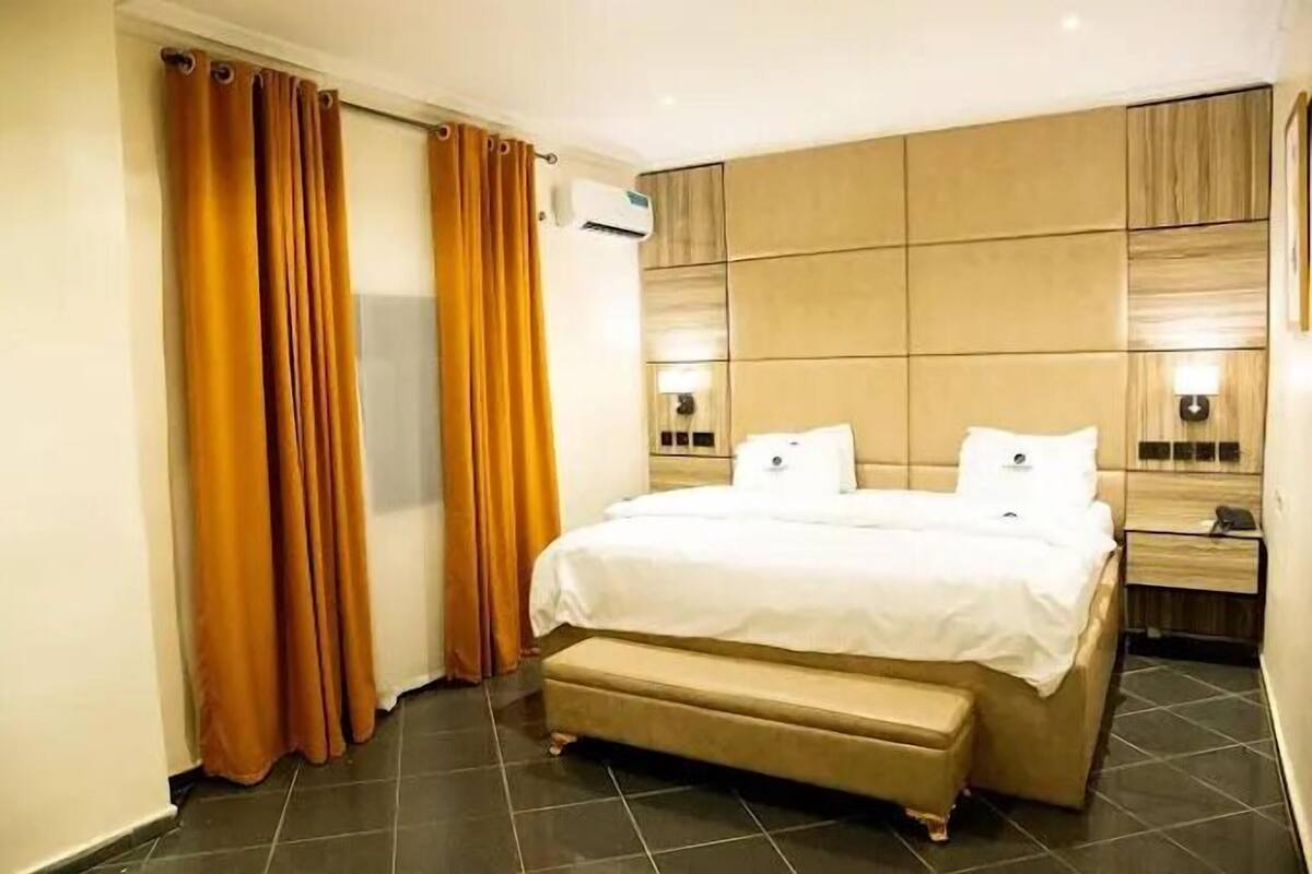 A hotel room with a large bed, a bedside table, an air conditioning unit, and curtains.