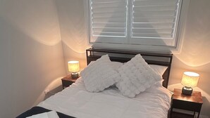 Una cama bien hecha con sábanas blancas y una colcha azul oscuro, dos almohadas blancas y toallas dobladas sobre la cama. Hay dos mesitas de noche con lámparas y una ventana con persianas al fondo.