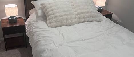 A neatly made bed with white bedding and fluffy pillows, flanked by two bedside tables with lamps, in a room with a window and a plain wall.