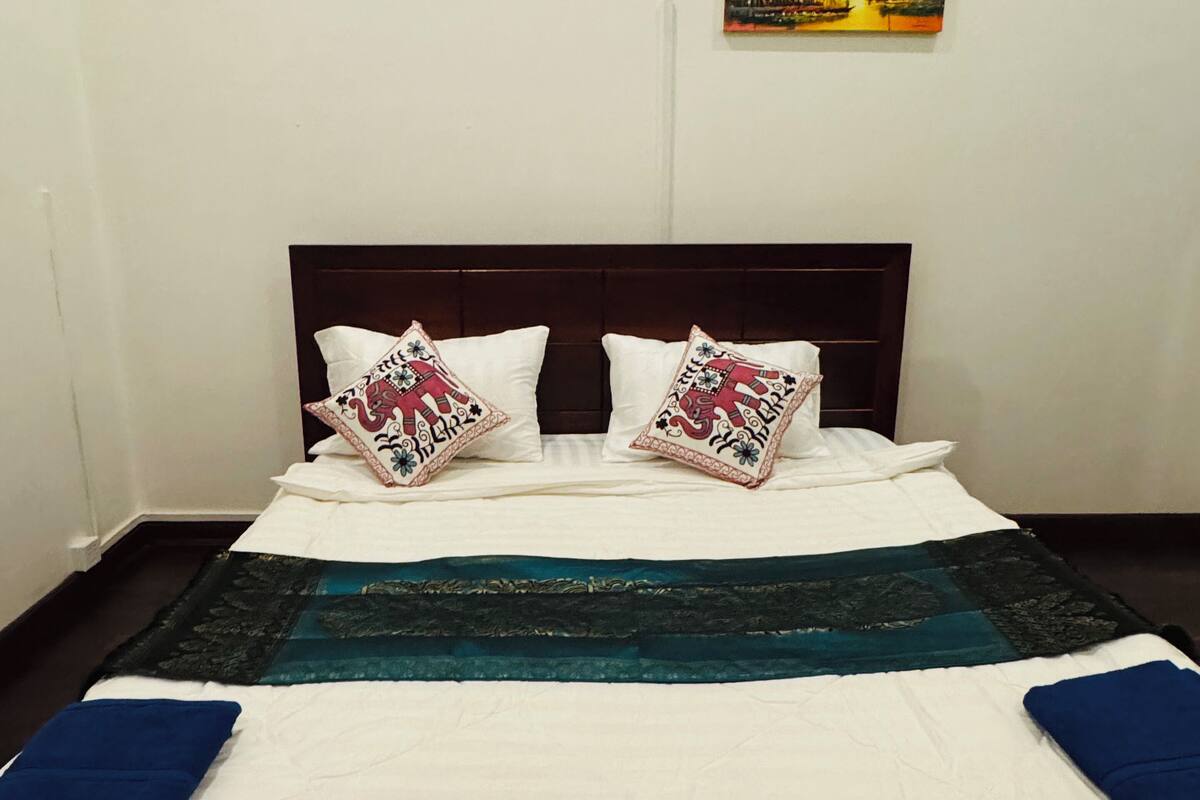 A neatly made bed with a white duvet, two decorative pillows, a dark headboard, and a painting of a sunset scene on the wall.