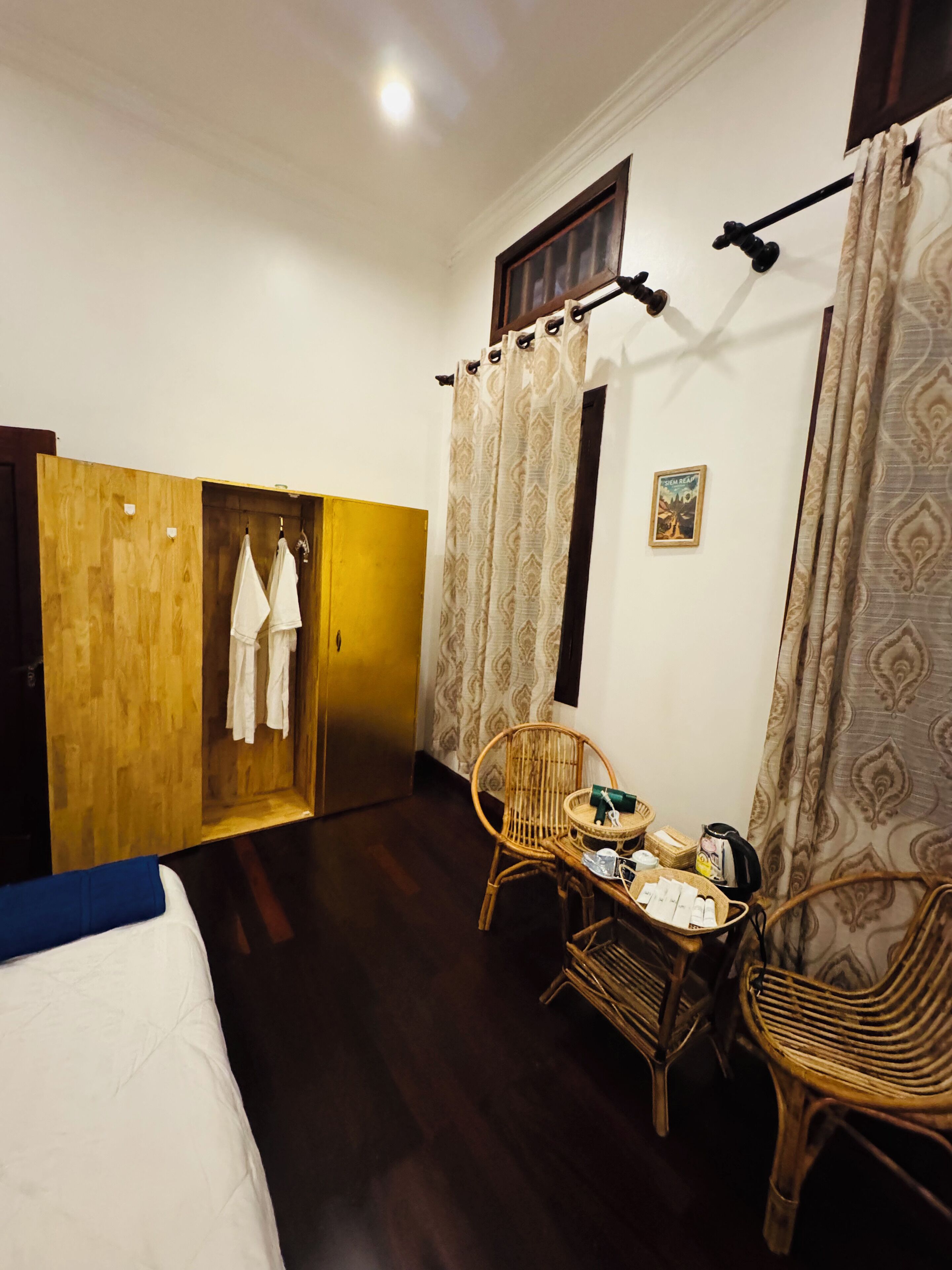 A hotel room with a bed, a wooden wardrobe, a small table with a chair, and a window with curtains.
