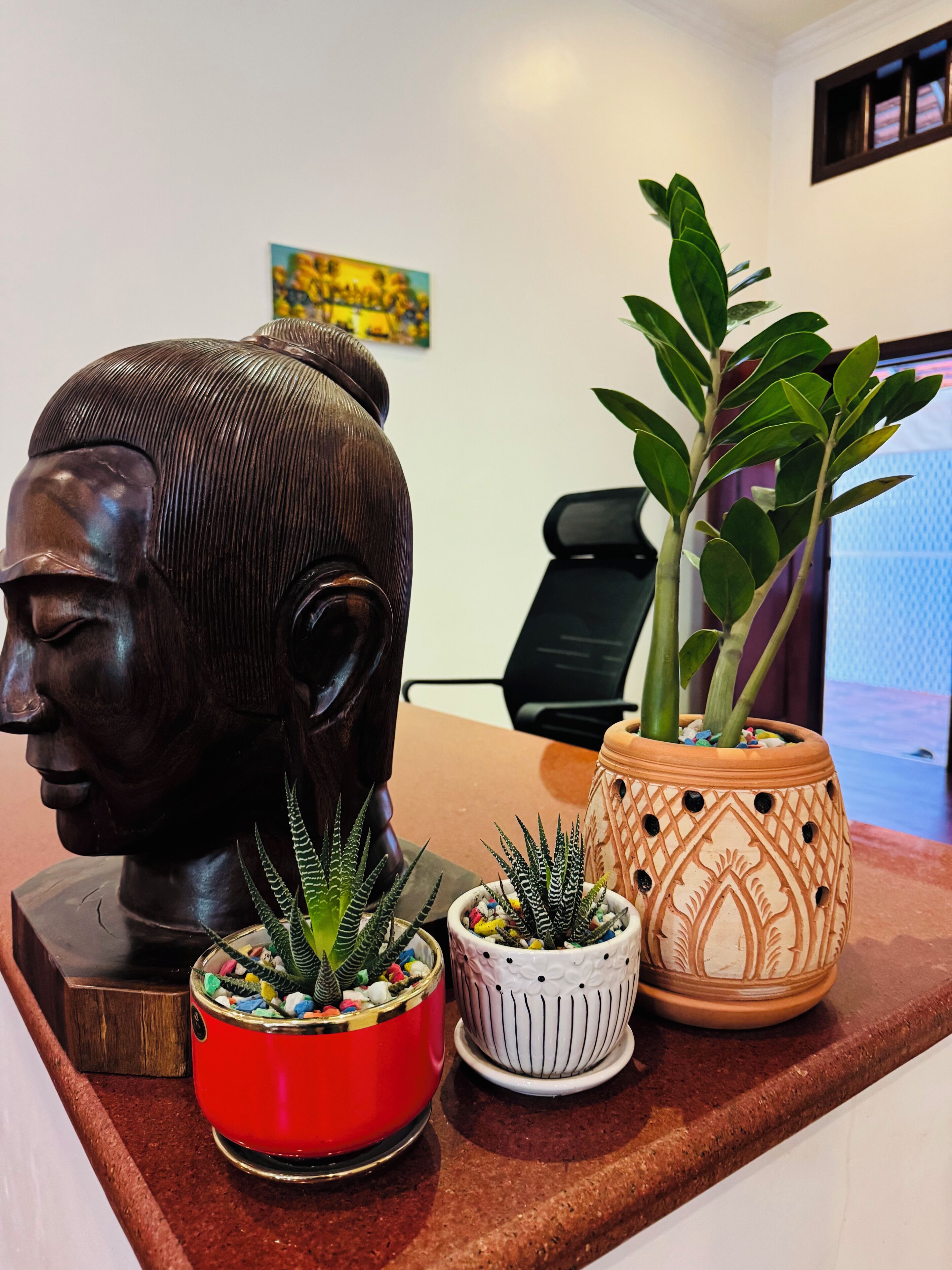 A bust sculpture, three potted plants, and a chair in a room with a white wall and a painting.