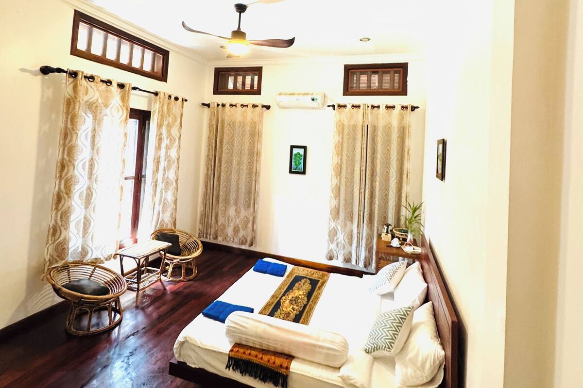 A room with a bed, two chairs, a ceiling fan, and a window with curtains.