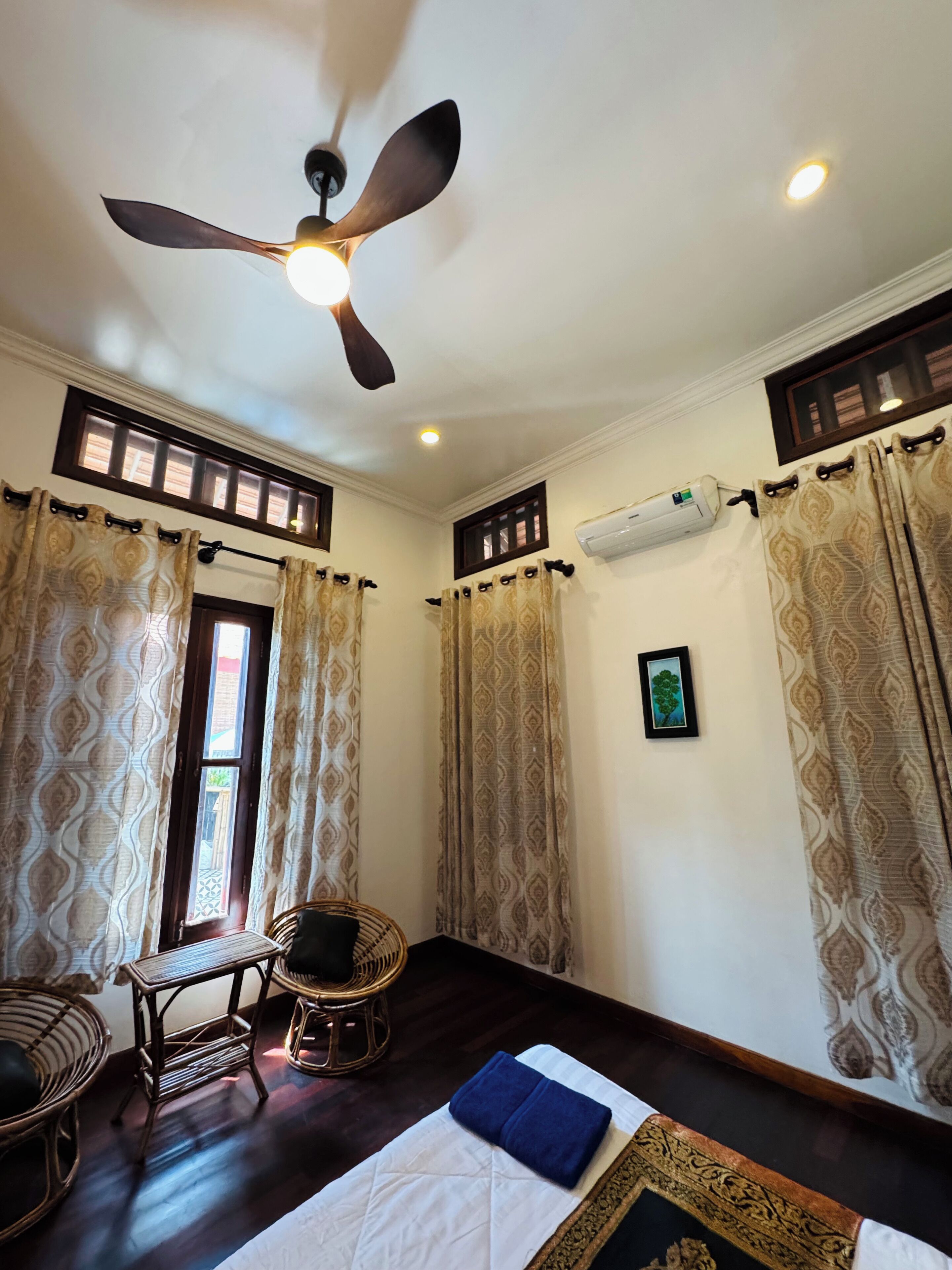 A room with a ceiling fan, patterned curtains, a bed with a blue pillow, wicker furniture, and an air conditioning unit.