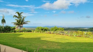 A grassy outdoor area with a concrete path, palm trees, and a view of distant hills and a body of water. - Doug's Place - Spectacular Views + Tennis Court (Nindaroo)