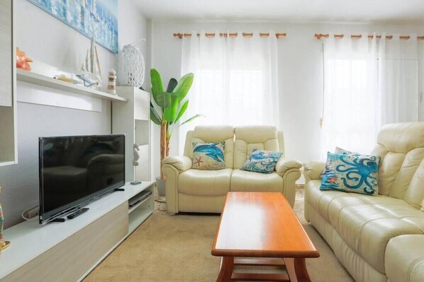 A living room with a beige sofa, a wooden coffee table, a TV on a stand, and a plant.