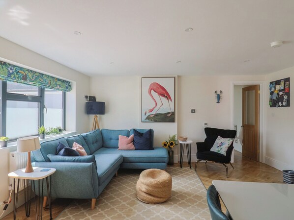 A living room with a blue sofa, a black armchair, a woven pouf, and a painting of a flamingo.