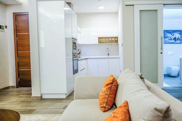 A modern living room with a sofa and orange cushions, a kitchen area with white cabinets, and a bathroom visible through an open door.