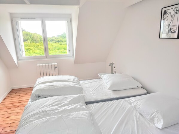 A bedroom with two beds, a window with a view of greenery, a radiator, and a framed picture on the wall.