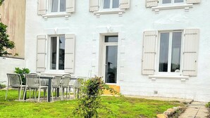 A white two-story house with multiple windows and shutters, a small lawn with a table and chairs, and a paved pathway. - LANDES HOLIDAY HOME (Penmarch)
