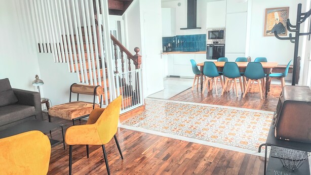 A modern living room with a wooden floor, a staircase, a dining area with blue chairs, and a kitchen with blue cabinets. - LANDES HOLIDAY HOME (Penmarch)