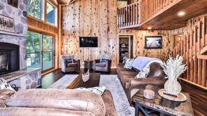 Interior acogedor de una cabaña con chimenea de piedra, paredes de madera, televisor, sofás de cuero y un balcón con barandilla de madera.