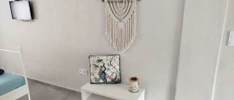 A minimalist room with a white bed, a small white bench, a macrame wall hanging, and a television mounted on the wall.