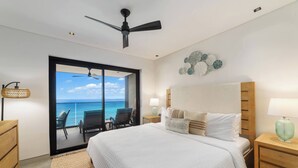 A bedroom with a large bed, a ceiling fan, a nightstand with a lamp, and a view of the ocean through a sliding door.