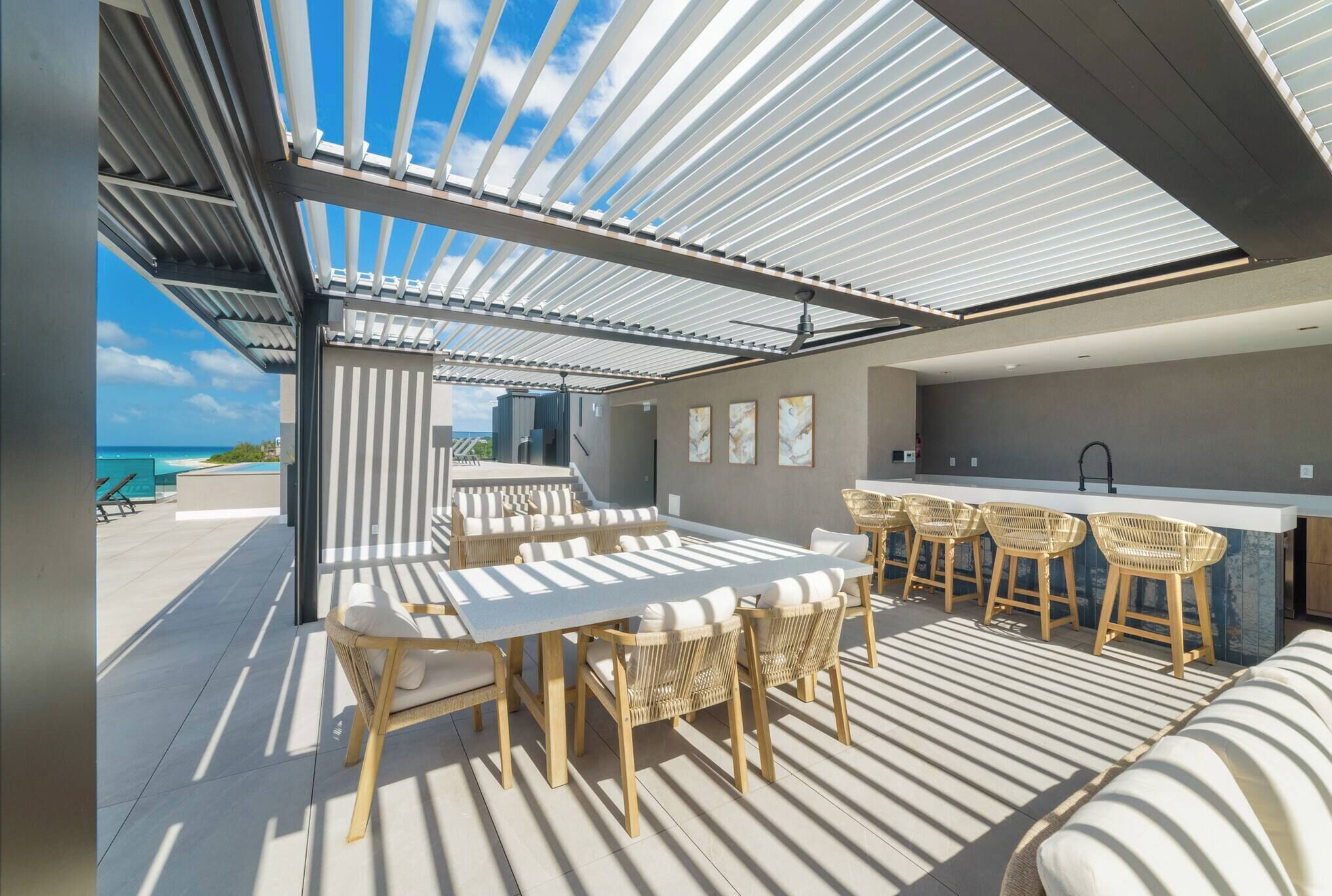 A modern outdoor dining area with a pool view, featuring a long table, several chairs, and a bar area.