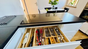 A kitchen with a black countertop, a drawer with utensils, a window with flowers, and a dining table with chairs. - Casa de Rainer Tommerup (Tommerup)