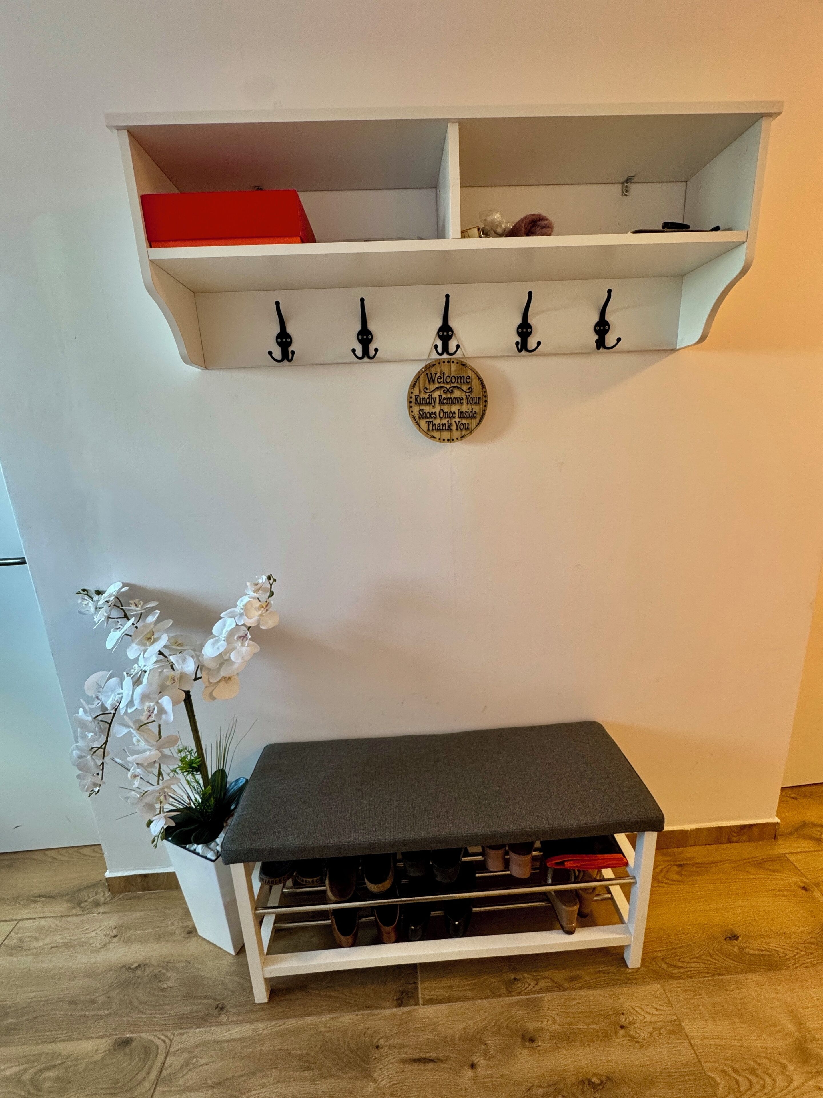 Una habitación con pared blanca, un banco de madera con espacio para guardar zapatos debajo, un estante blanco pequeño con una caja roja, y un cartel en la pared sobre el banco.
