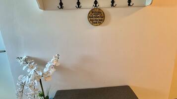 Una habitación con pared blanca, un banco de madera con espacio para guardar zapatos debajo, un estante blanco pequeño con una caja roja, y un cartel en la pared sobre el banco.