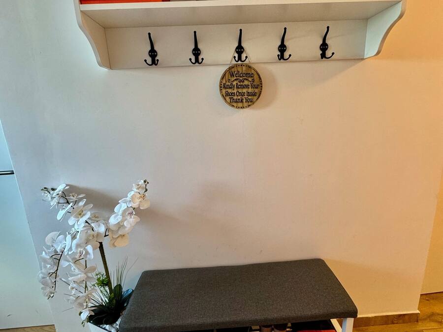 Una habitación con pared blanca, un banco de madera con espacio para guardar zapatos debajo, un estante blanco pequeño con una caja roja, y un cartel en la pared sobre el banco.