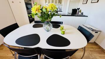 A dining table with a vase of yellow flowers, two green candles, and black chairs in a modern kitchen with white cabinets and a built-in oven.