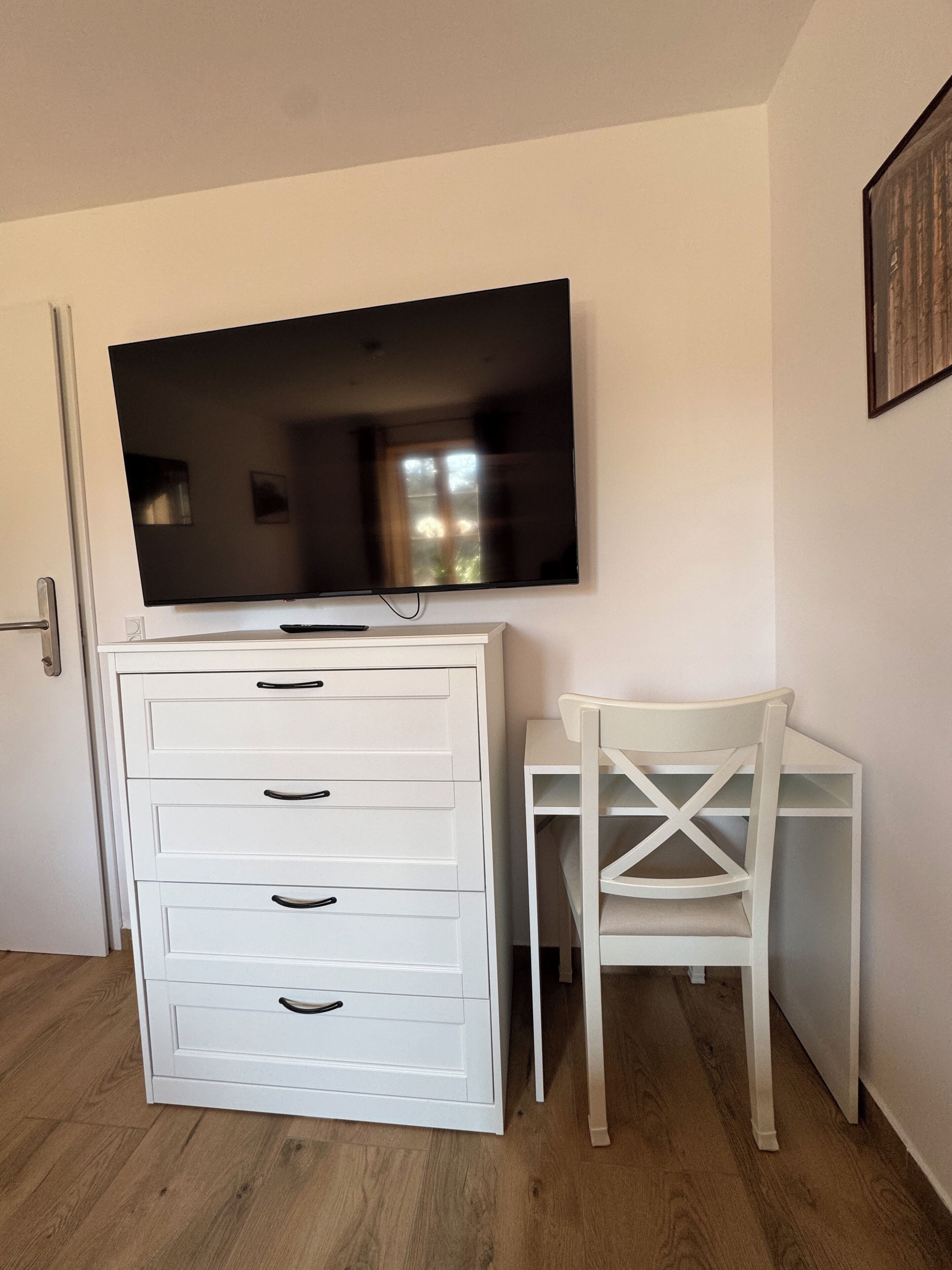 Una habitación con una televisión de pantalla plana montada en la pared, un aparador blanco con cajones y una silla blanca con respaldo de cruces.