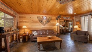 Interior acolhedor de cabana com teto de madeira, sofá de couro marrom, mesa de centro de madeira e um espaço de jantar com mesa e cadeiras.