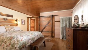 Quarto com teto de madeira, cama com colcha floral, um banco de madeira, cômoda com espelho e porta de celeiro deslizante.