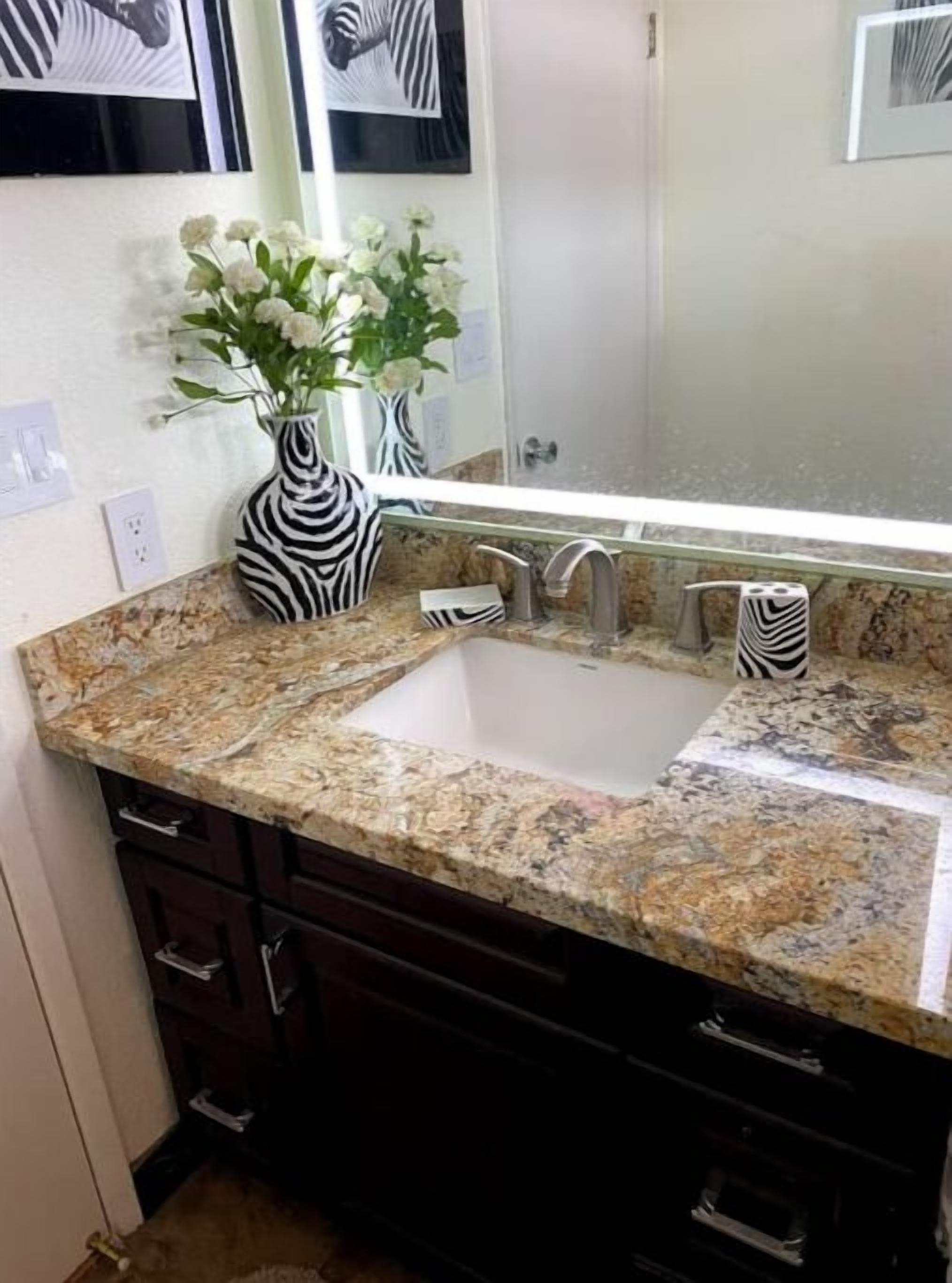 Un baño con encimera de granito, un lavabo blanco y un espejo encima. Hay dos jarrones con flores blancas sobre la encimera.
