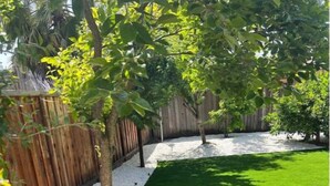 Un patio con césped artificial, valla de madera y un árbol proyectando sombras.