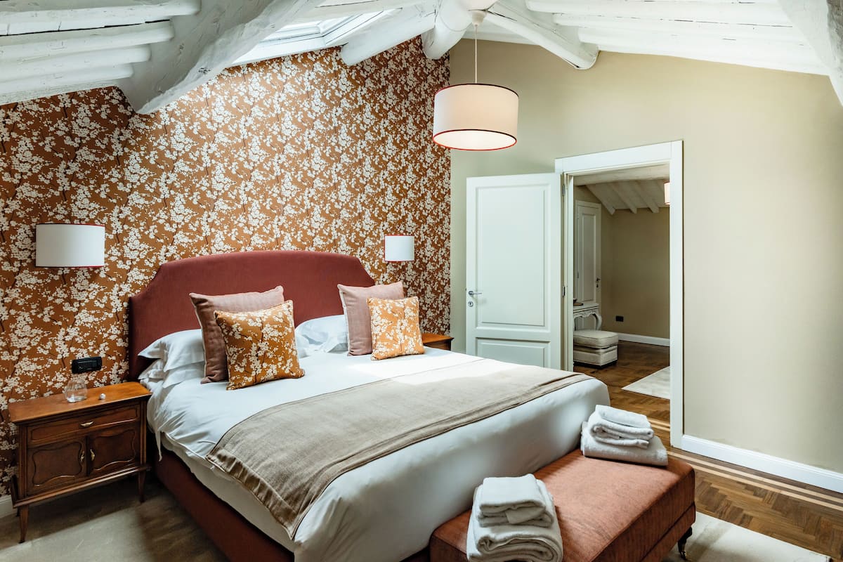 A bedroom with a floral wallpaper, a red headboard, a bed with white and orange pillows, a bedside table, and a hanging light fixture.