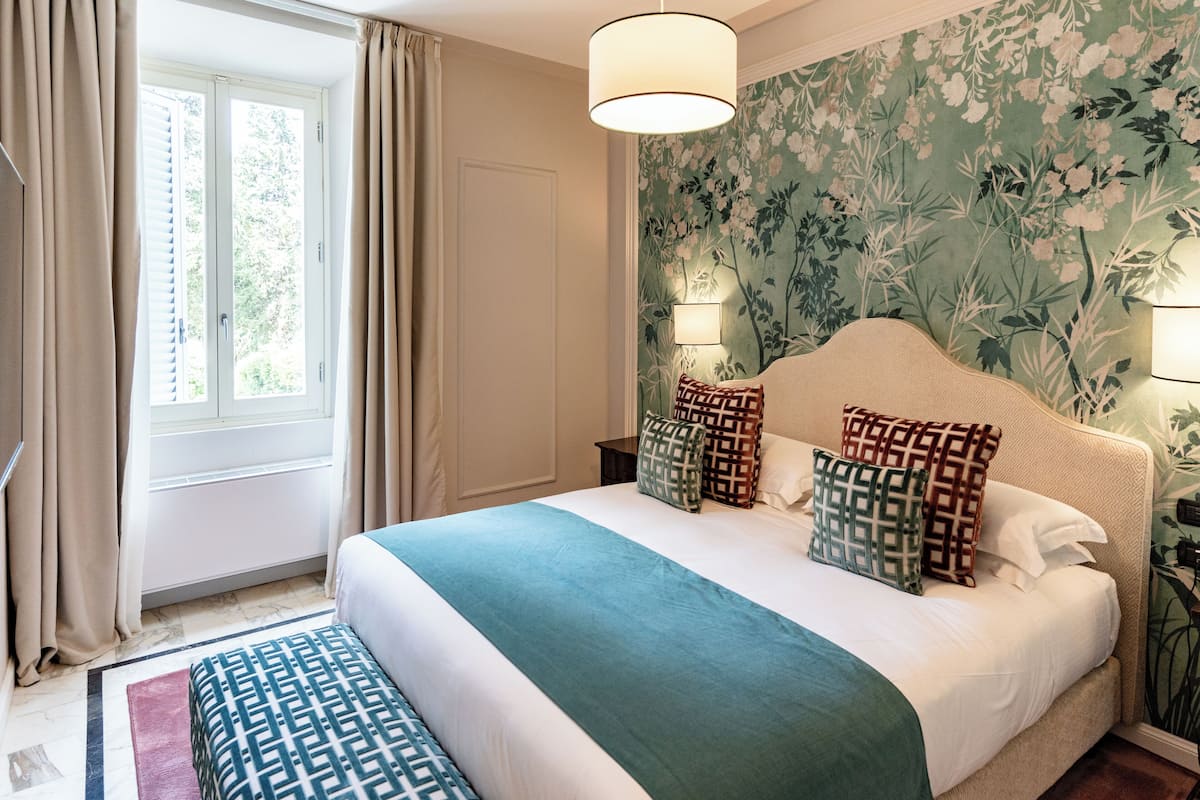 A bedroom with a large bed, a window with curtains, a headboard, and a floral wallpaper.