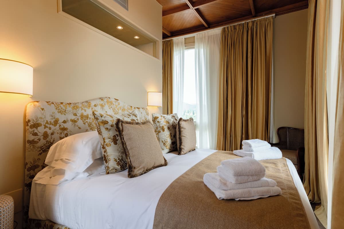 A hotel room with a large bed, floral headboard, and a view of the outdoors through a window with curtains.