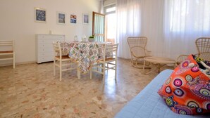 A room with a dining table, chairs, a bed, a dresser, and a window with curtains.