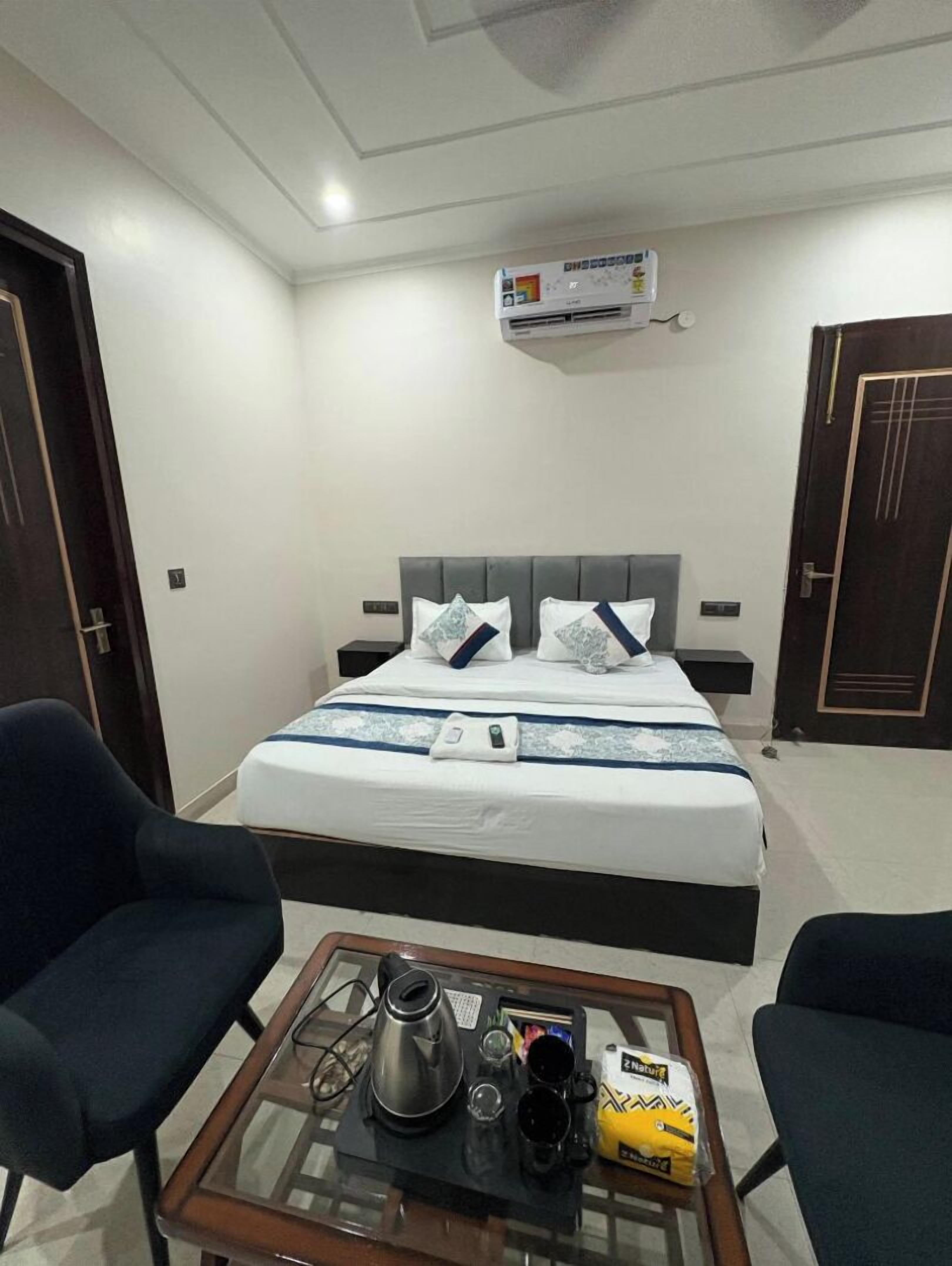 Habitación de hotel con cama, dos sillas, una mesita con hervidor, tazas y un paquete de aperitivos.