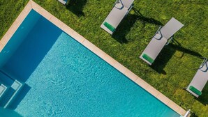 An aerial view of a rectangular swimming pool with a tiled border, surrounded by a well-maintained lawn and three lounge chairs with green cushions.
