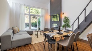 A modern living room with a grey sofa, a dining table with chairs, a TV mounted on the wall, and a staircase.