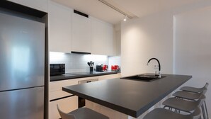 A modern kitchen with a stainless steel refrigerator, white cabinets, a black countertop, a red kettle, and a bar stool area.