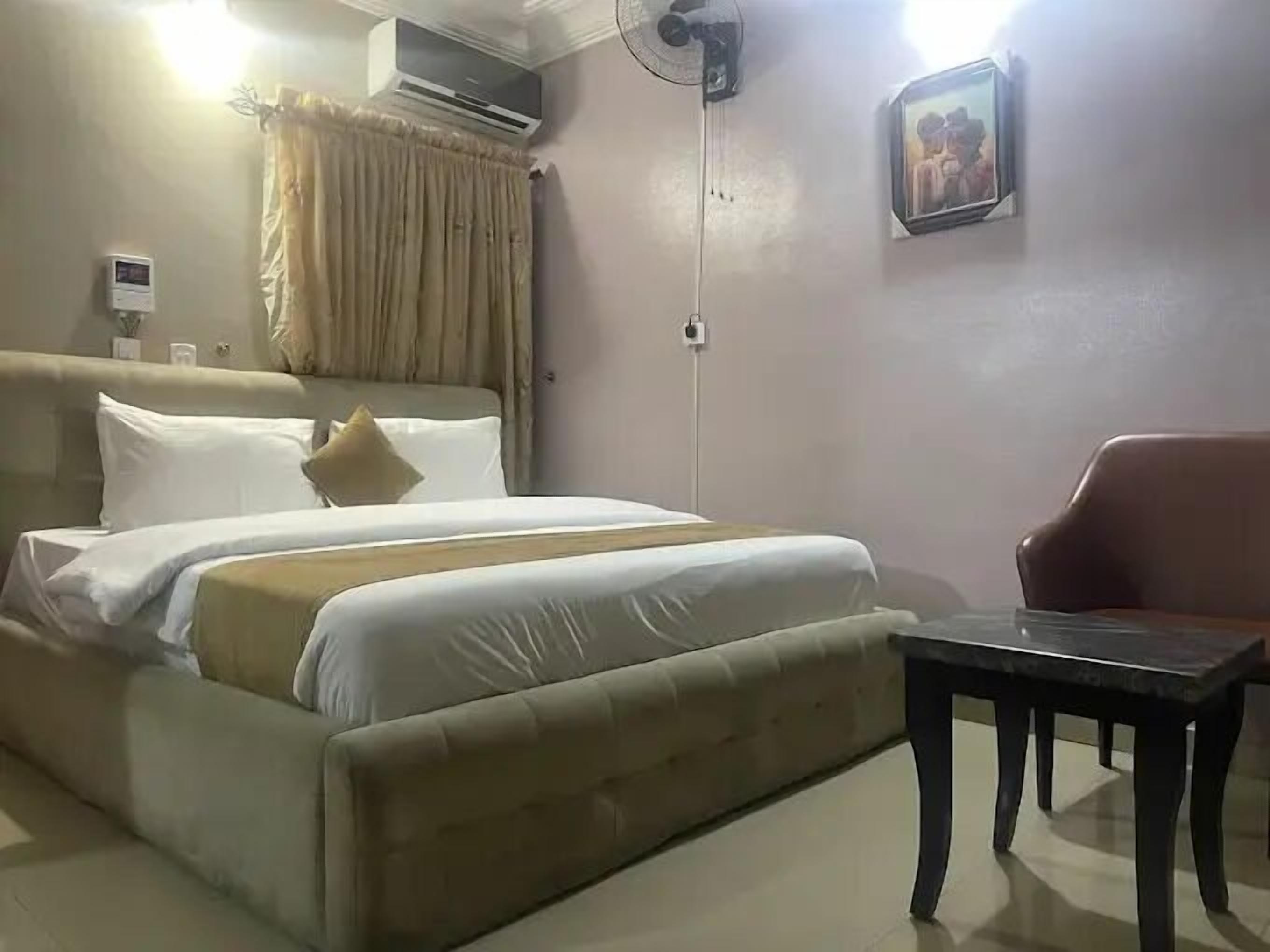 Een hotelkamer met een bed, een stoel, een klein tafeltje, een airconditioner, een ventilator en een schilderij aan de muur.