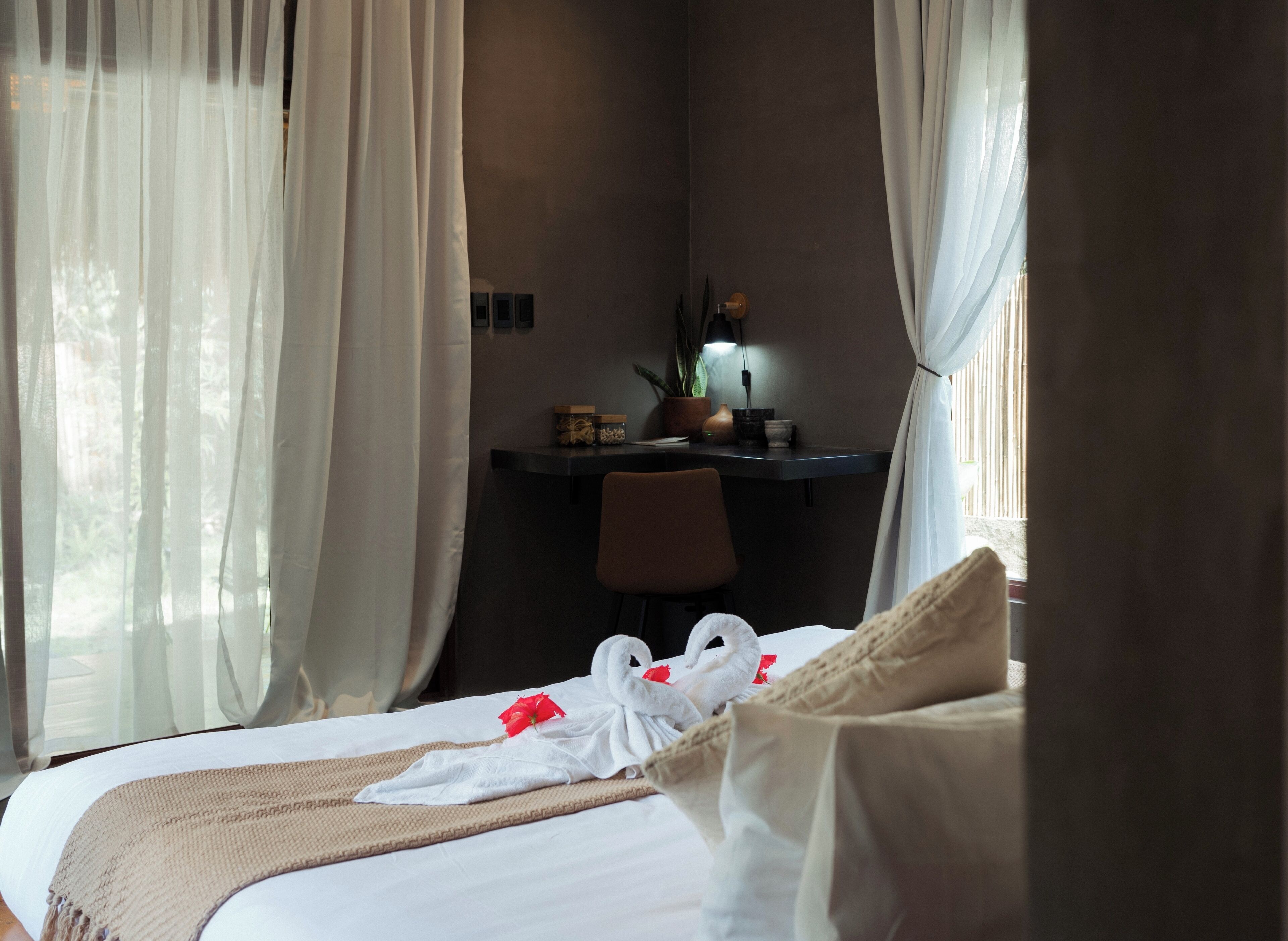 A hotel room with a bed, a desk, and a chair. There are curtains on the windows, and a plant is on the desk.