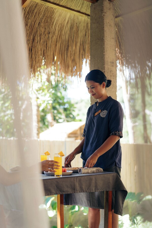 A person preparing food at an outdoor table with a thatched roof, surrounded by greenery.
