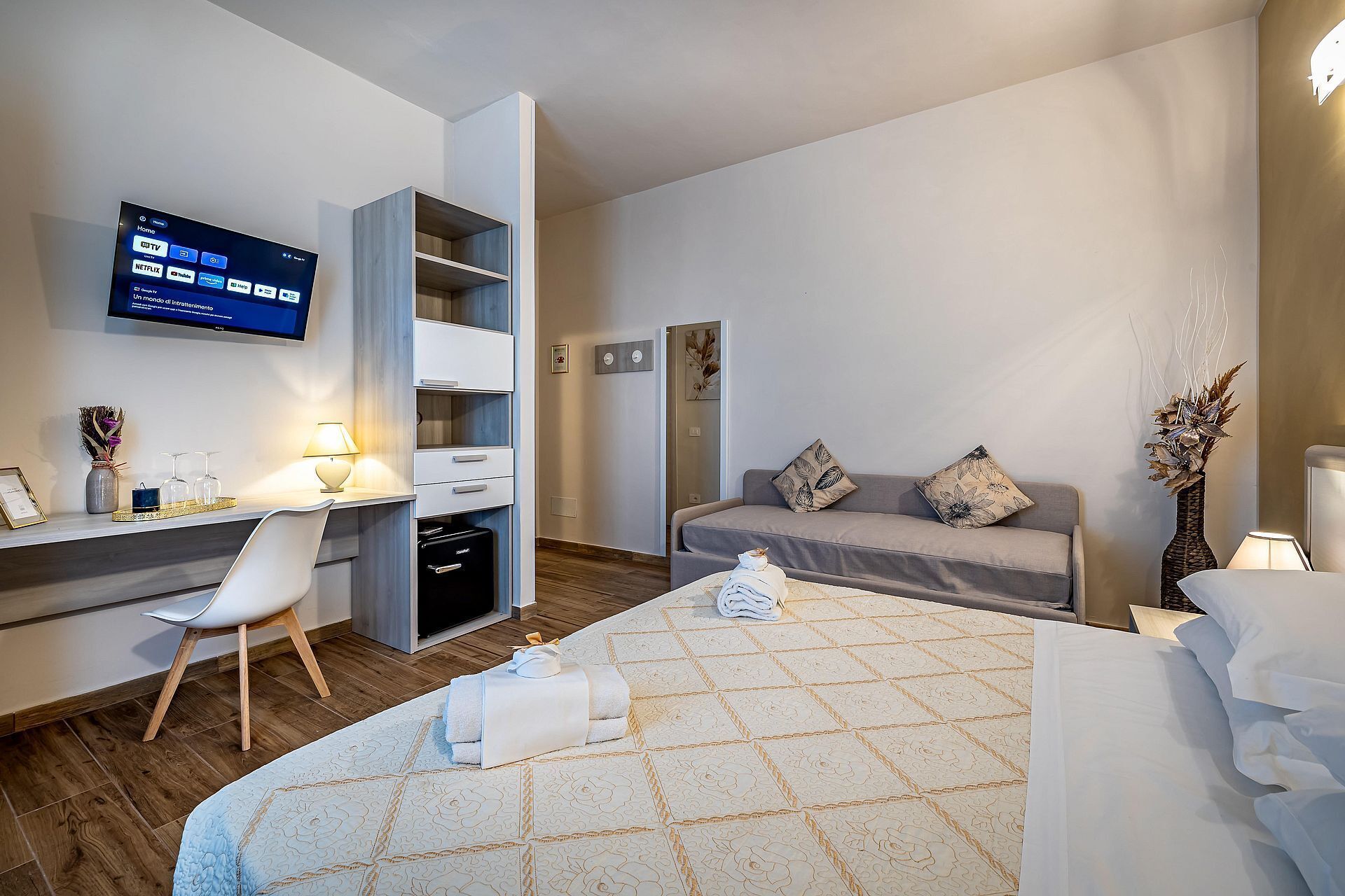 A modern hotel room with a bed, a desk with a computer, a TV, and a sofa.