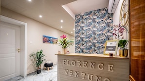 A reception desk with "FLORENCE VENTUNO" signage, a floral wall, a cityscape painting, and potted plants.