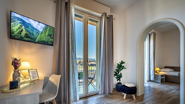 A modern living room with a large TV, a dining area with a table and chairs, a balcony with a view of the mountains, and a cozy sitting area with a bed.
