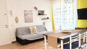 Uma sala de estar com sofá, mesa de jantar com cadeiras, televisão e janela com cortinas.