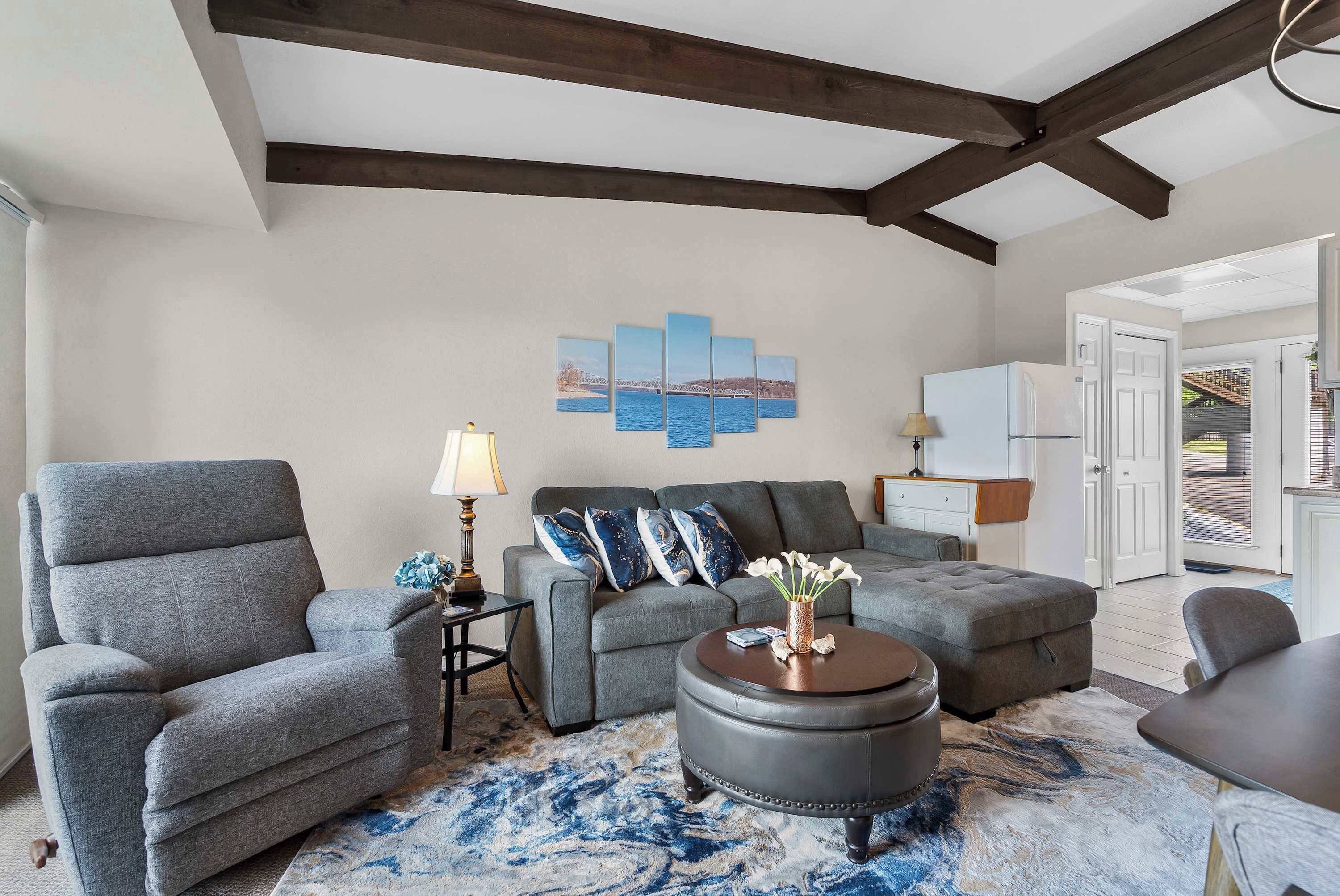 Un salon avec un canapé gris, un pouf rond, une table basse et une œuvre d’art accrochée au mur.