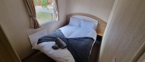A small, neatly made bed with a white headboard, a grey blanket, and a rolled-up towel.