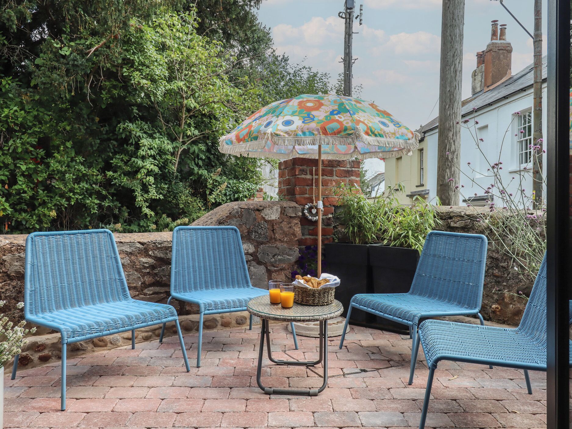 A patio with three blue wicker chairs, a round table with a basket of fruit and a bottle of orange juice, and a colorful umbrella.